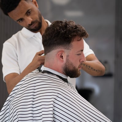 Side View of a customer having his hair cut by a barber while sat in a striped gown