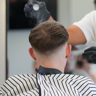 Barber putting styling powder on cutomers hair while he is relaxed in a barber chair with a cape