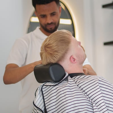 Barber cutting a ginger customers hair while he is relaxed in a barber chair with a cape