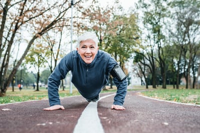 a woman with short white hair and running clothes is doing a push up outside in a park path