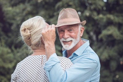 An older man in a fedora and white beard and a stylish older woman are embracing