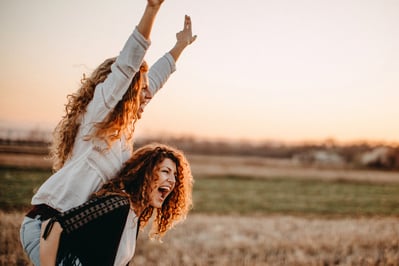 two joyful women with curly long  hair are celebrating, one lifting the other up on her shoulders 