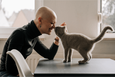 a smiling woman in a black turtle neck and a  bald head and a grey cat are touching nose to nose