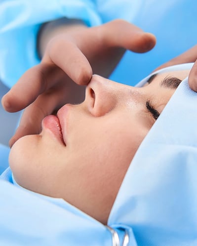 Patient draped in blue, surgeon's hands examine nose for rhinoplasty.