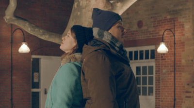 A young woman and man in winter coats stand with their backs together.