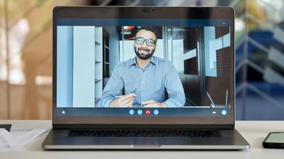 A laptop screen showing a video call with a man in a business shirt holding a pen