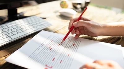 a person proofreading and correcting a document with a red pencil, computer in the background