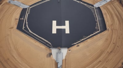 An aerial view of a circular helipad located in an open, grassy area. The helipad is marked with an 'H' in the center and surrounded by a circular path that has worn a darker ring in the grass. A straight path leads from the lower left toward the helipad. Shadows and sunlight create interesting textures on the ground.