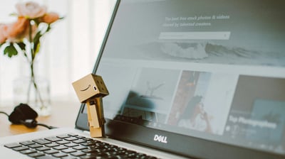 computador aberto com clip segurando uma caixa com logo amazon um vaso de flores amarelas ao fundo