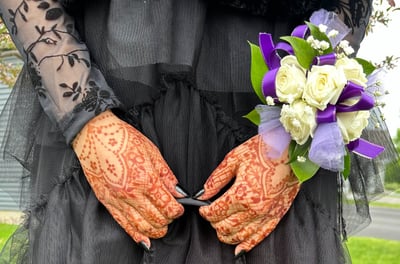 Close up of two hands with henna on them, holding a purse strap, wearing a black dress & corsage