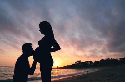Um casal, sendo ela, grávida, e ele beijando sua barriga, numa praia deserta, ao por do sol.
