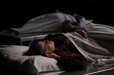 21 North - Two actors laying on beds, one in the foreground and one in the background out of focus.