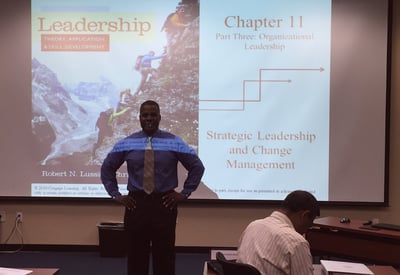 Dr. Marlon standing in blue shirt, light-colored tie and dark pants in front of a projector screen