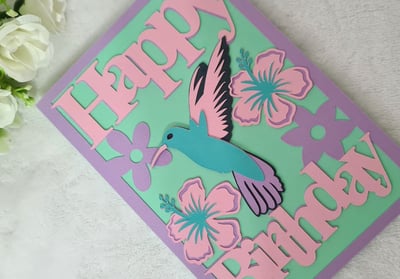 A pastel-themed birthday card with "Happy Birthday" text, featuring a vibrant blue hummingbird and p
