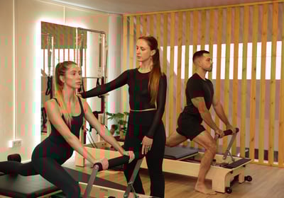 Pilates instructor guiding a student on a duo reformer, while another person exercises in the back
