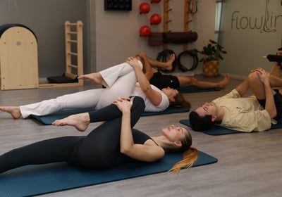 People lying on mats in a Pilates studio, stretching with a single leg stretch pose, beginner level.