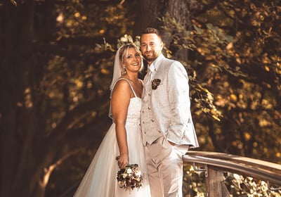 Brautpaar in eleganter Hochzeitskleidung auf einer Holzbrücke umgeben von Bäumen