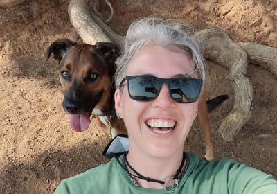 A smiling person in sunglasses takes a selfie under a tree with a dog seated smiling behind them