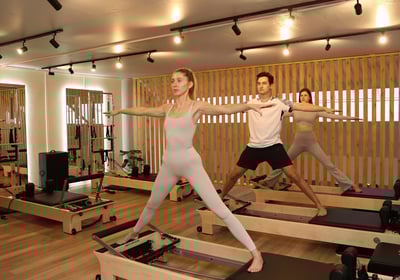Três pessoas praticando Pilates em reformers, seguindo a instrutora na posição Guerreiro II.