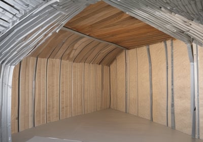 A house is under construction with visible wooden framing and scaffolding around the roof area. The building is enclosed by a temporary metal fence, and there are materials and construction tools placed around the site. The roofing is partially complete, made of dark grey tiles, and the surrounding area includes grass and a paved sidewalk.