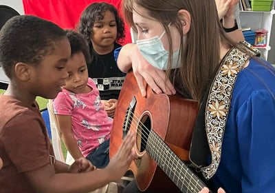 children surrounding woman with guitar and a child touching guitar strings