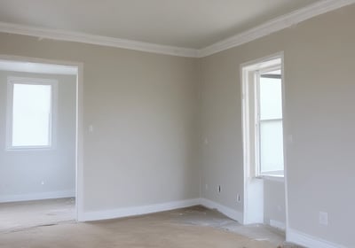 A partially constructed or renovated interior space featuring exposed brick walls and wooden ceilings. A white door is visible in the background next to a red painted wall. A plastic sheet covers furniture on the right side, and there are construction materials scattered across the floor.