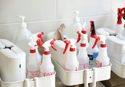 Estante de baño con varios pulverizadores y botellas de limpieza de color blanco y etiquetas rojas