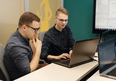 Dois adultos em frente a um computador, sendo que um deles está ensinando o outro como operar
