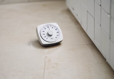 A bathroom scene featuring a vintage-style bronze bathtub faucet with a handheld shower attachment and flexible hose, set against a white bathtub. The background includes a wooden surface with decorative items like a round dish and textured white vase. Natural light streams in from a nearby window, casting soft shadows.