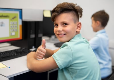Smiling student giving thumbs up while using a computer in a classroom
