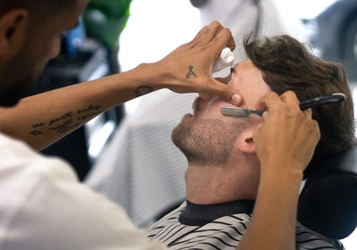 man receiving a shave with a cut throat razor by a barber while sitting in a chain in a barbershop