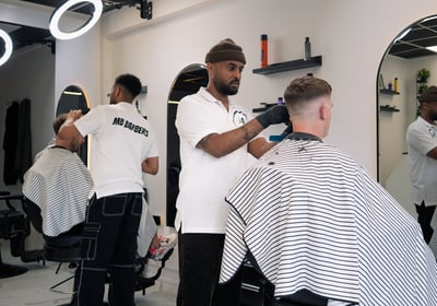 two barbers tending to cutting the hair of two customers while they are sitting in the chair