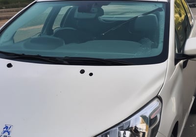 A close-up of a red car's front hood and windshield, featuring black wiper blades. The windshield reflects the sky and surrounding environment, while a pack of gum is visible on the dashboard. The car's surface is shiny, capturing subtle reflections.