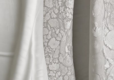A white lace wedding or bridal dress is hanging on a black hanger in front of a blue trash bin. The dress includes intricate lace details and layered fabric. A tag with an image of a model wearing a similar garment is attached to the hanger.