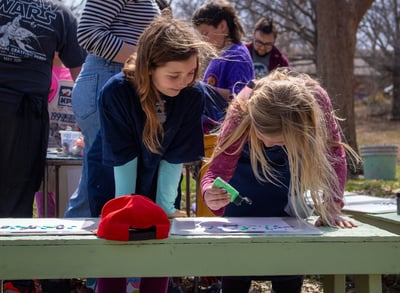 Two young girls smiling while standing over papers dripping paint onto them.