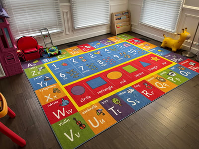 Photo of daycare room where children play. Large colorful floor mat and bookshelf.