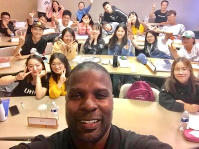 Dr. Marlon Ware standing in front of his class with students holding up the peace sign