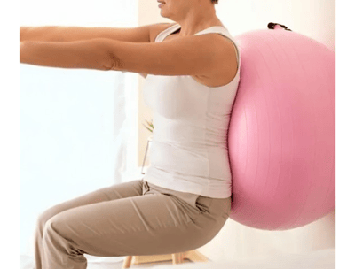 Paciente encostada na parede com uma bola rosa de pilates nas costas realizando exercício