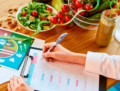 Mãos de nutricionista escrevendo em um prontuário sobre uma mesa com alimentos saudáveis