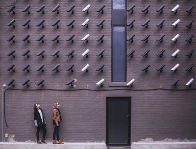 Two people standing in front of a brick wall that has many video surveillance cameras.