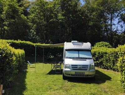 Spacious campervan pitch on grass with awning and outdoor table, surrounded by hedges and forest