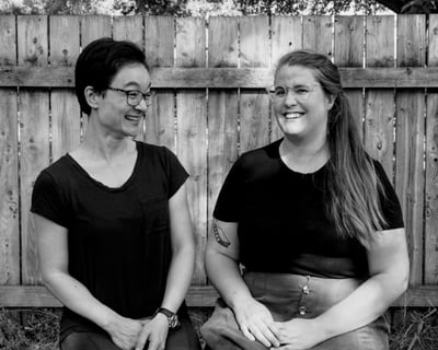 Andy and Jen laughing together while seated outdoors in dappled sunlight in front of a fence.