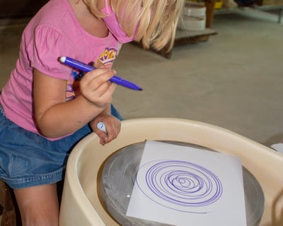 Young girl sitting at a potters wheel holding a marker, making spin art.