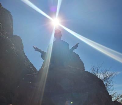 The silhouette of a person on a mountain meditating with the sun in the background.