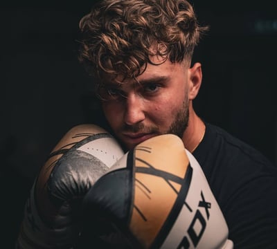 Barbat cu manusi de box RDX, in garda, priveste concentrat spre camera.