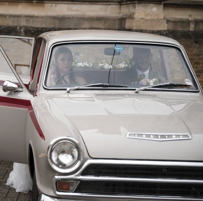 Bride and groom sit in a vintage Ford Cortina wedding car with an open door.
