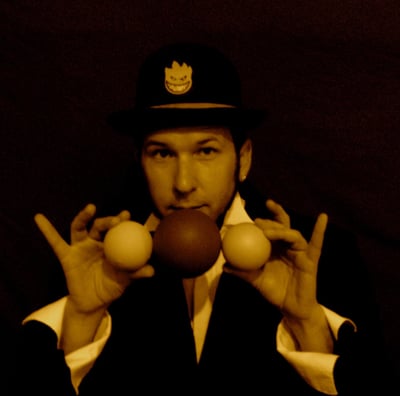 a man in a suit and hat with two hands up to the camera holding juggling balls
