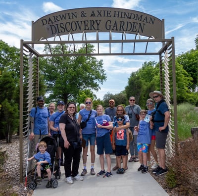 Group photo of adults and children posed on a paved walkway under an entry to a garden