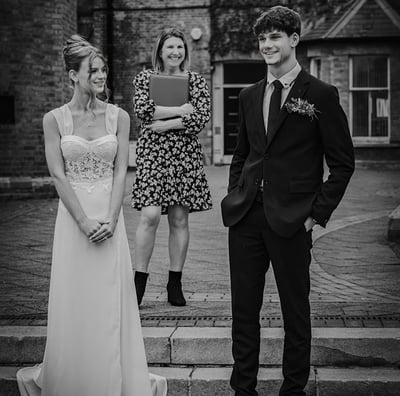 A black & white photo of a bride & groom smiling at each other on steps, officiant behind them, in front of a brick building.