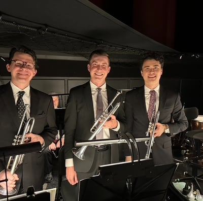 Three men holding trumpets and wearing suits, in an orchestra pit.